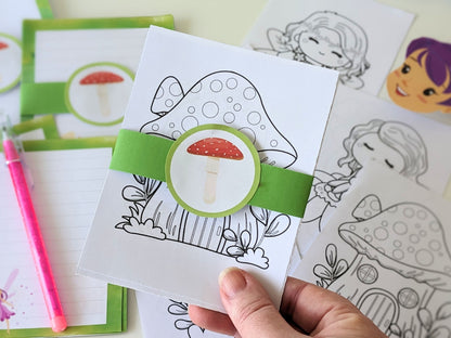 Coloring book with fairy mushroom design held by a hand, surrounded by other coloring pages.