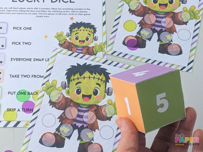 A close-up of a hand holding a colorful paper game die in front of printed game cards featuring a cartoon Frankenstein character. The scene shows the product being used, with a visible hand and printed board/game materials in a real setting, so this fits Lifestyle rather than Product/Packaging.